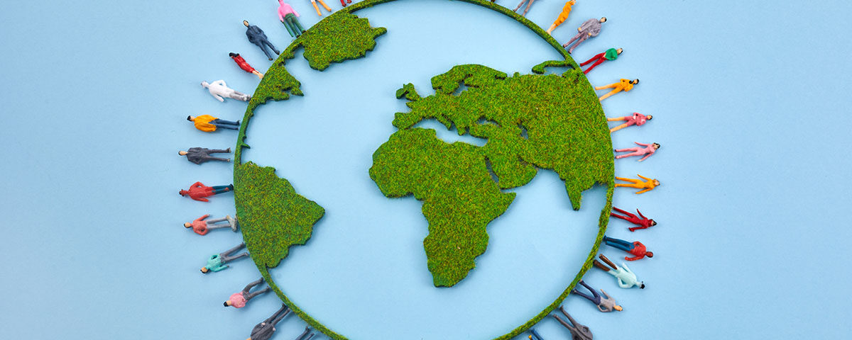 Representação estilizada do globo terrestre visto de cima, com os continentes (destacando-se África, Europa e Américas) formados por uma textura verde que lembra musgo ou grama. Ao redor do globo, um círculo de diversas miniaturas de figuras humanas coloridas e variadas estão de pé, como se estivessem sobre a borda do planeta, todas voltadas para fora ou em diferentes direções. O fundo é de um azul claro uniforme. A imagem evoca a ideia de responsabilidade coletiva global e a interdependência entre a humanidade e o meio ambiente, contextualizando a necessidade e a importância das leis ambientais, como as principais leis do Brasil, para a proteção do planeta e a regulação das atividades humanas em prol da sustentabilidade.