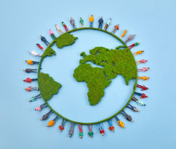 Representação estilizada do globo terrestre visto de cima, com os continentes (destacando-se África, Europa e Américas) formados por uma textura verde que lembra musgo ou grama. Ao redor do globo, um círculo de diversas miniaturas de figuras humanas coloridas e variadas estão de pé, como se estivessem sobre a borda do planeta, todas voltadas para fora ou em diferentes direções. O fundo é de um azul claro uniforme. A imagem evoca a ideia de responsabilidade coletiva global e a interdependência entre a humanidade e o meio ambiente, contextualizando a necessidade e a importância das leis ambientais, como as principais leis do Brasil, para a proteção do planeta e a regulação das atividades humanas em prol da sustentabilidade.