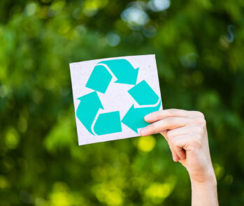 Mão de uma pessoa segura um pequeno cartão quadrado branco, onde está impresso o símbolo universal da reciclagem (três setas verdes formando um ciclo) em destaque. O fundo é composto por folhagem verde e desfocada, sugerindo um ambiente natural e realçando a mensagem sobre a importância da reciclagem para a sustentabilidade e a preservação do meio ambiente.