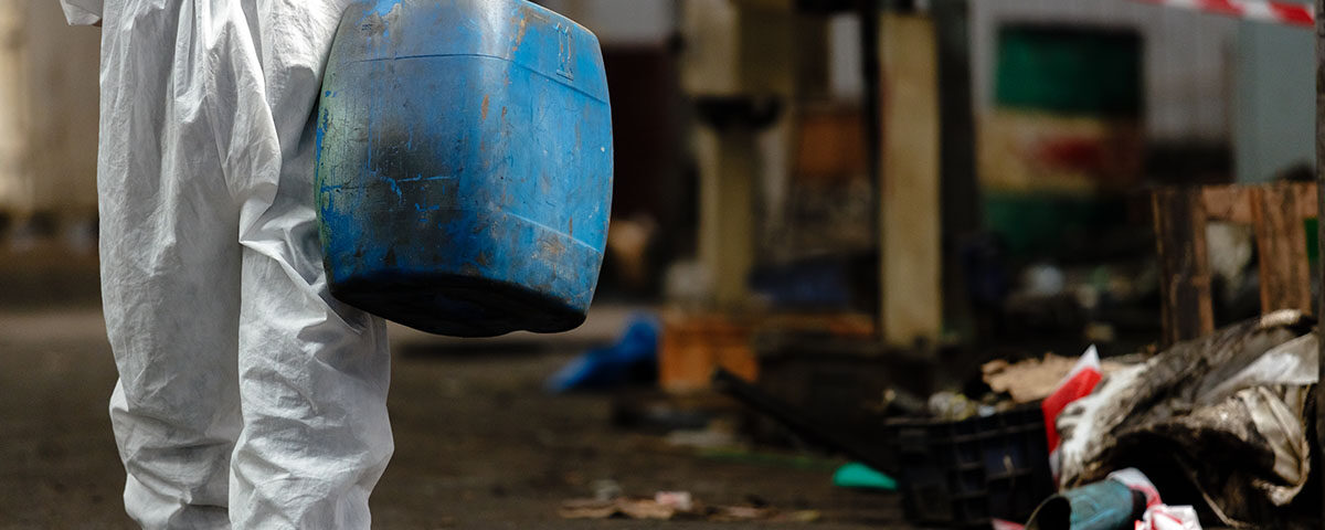 Pessoa vista parcialmente por trás, trajando um macacão de proteção branco completo que cobre todo o corpo e sapatos, enquanto carrega um grande tambor (ou bombona) de plástico azul, aparentemente pesado. O cenário é um ambiente industrial ou de descarte, com piso escuro, alguns detritos e uma fita de isolamento vermelha e branca ao fundo, sugerindo uma área de acesso restrito ou manuseio de materiais específicos. A imagem ilustra o processo de manejo de resíduos que exigem equipamentos de proteção individual (EPIs), possivelmente resíduos de classe IIA ou IIB, conforme o tema.
