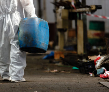 Pessoa vista parcialmente por trás, trajando um macacão de proteção branco completo que cobre todo o corpo e sapatos, enquanto carrega um grande tambor (ou bombona) de plástico azul, aparentemente pesado. O cenário é um ambiente industrial ou de descarte, com piso escuro, alguns detritos e uma fita de isolamento vermelha e branca ao fundo, sugerindo uma área de acesso restrito ou manuseio de materiais específicos. A imagem ilustra o processo de manejo de resíduos que exigem equipamentos de proteção individual (EPIs), possivelmente resíduos de classe IIA ou IIB, conforme o tema.